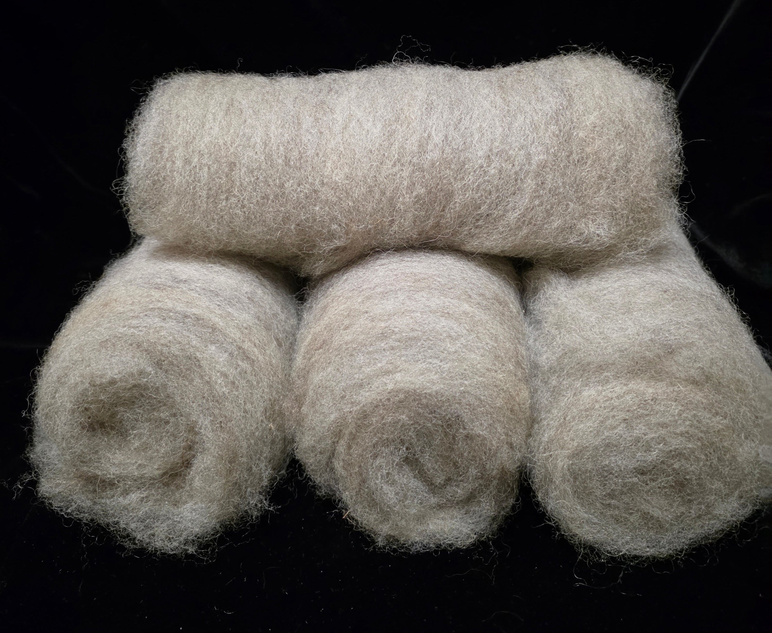 Four rolled batts of fluffy grey romney wool stacked on a solid black background.