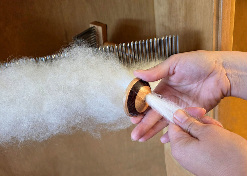 A close-up of a person's hands using a diz to pull combed wool off of a hackle into a long stream of hand combed top set against a wooden background. 